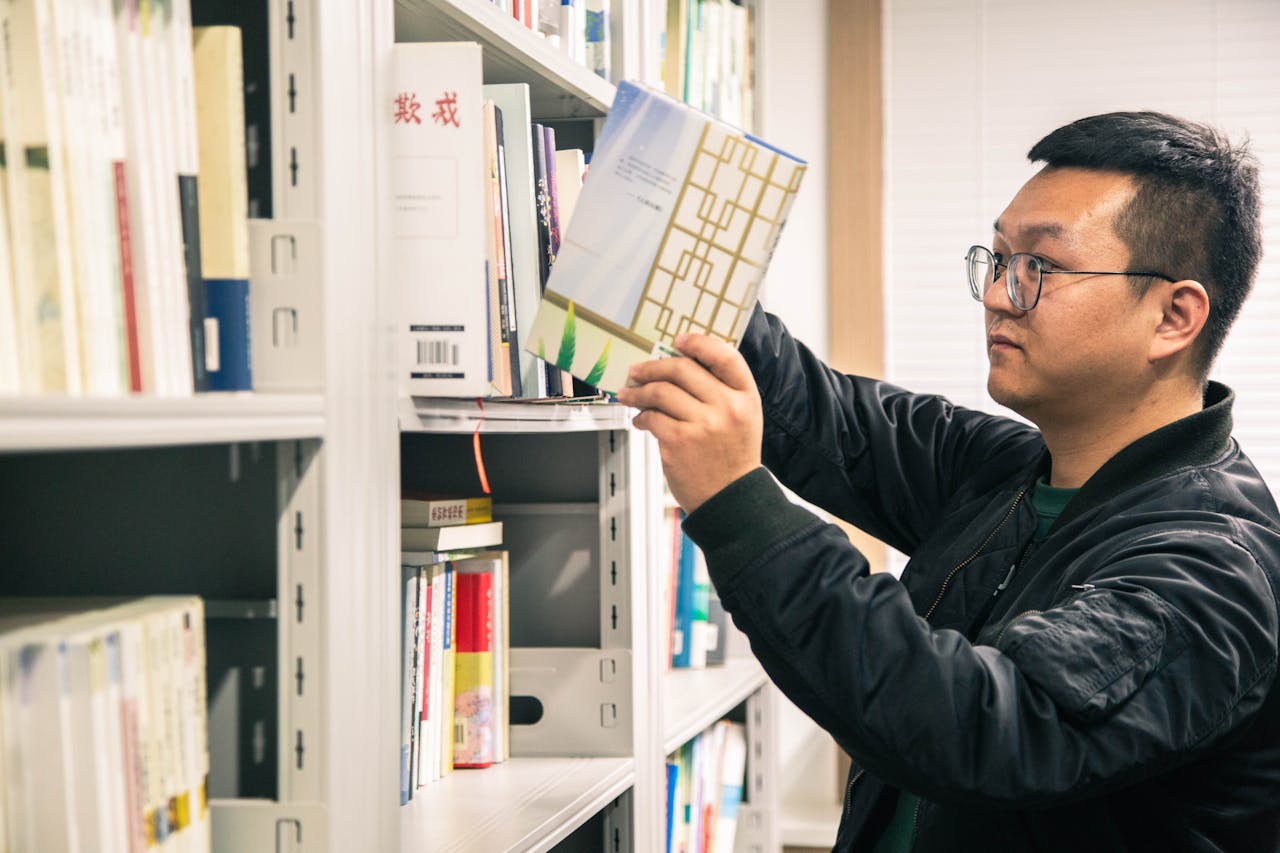 portfolio-01 Asian man in glasses selecting a book from a library shelf, focused and thoughtful.